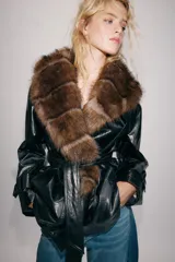 Trench corto Zara de efecto piel color negro, con cuello de solapa y aplicación de pelo sintético a contraste en color marrón. Cuenta con manga larga con trabillas en los puños, bolsillos delanteros y cinturón ajustable del mismo tejido.