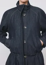 Parka oversize de tejido denim color azul oscuro, con cuello alto, mangas raglán largas, bolsillos laterales, cintura elástica y cierre frontal con botones ocultos.