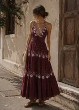 Vestido largo de estilo bohemio en color bordó, con escote profundo tipo halter y espalda descubierta. Presenta detalles de bordados florales blancos en el pecho y a lo largo de la falda, con diseño de paneles escalonados y cintura ajustada.