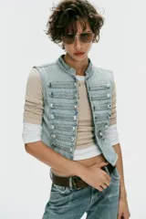 Chaleco de denim color celeste con cuello subido, sin mangas, con detalle de aplicaciones horizontales y botones metálicos en el frente. Cuenta con cierre frontal con ganchos metálicos ocultos.
