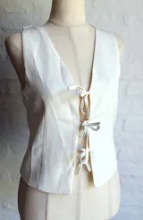 Chaleco blanco sin mangas, con escote en V profundo y cierre frontal mediante lazos anudados.