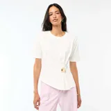 Camiseta blanca de manga corta y cuello redondo, con diseño drapeado en el lateral y detalle de adorno metálico dorado en la cintura.