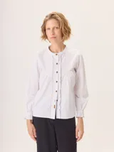 Camisa de algodón color blanco con cuello mao, cierre frontal con botones y detalle de volados en la tapeta. Presenta mangas largas con puños elásticos y diseño fluido.