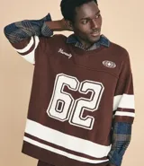 Remera oversized deportiva color marrón con cuello en V, rayas blancas en las mangas y en la parte inferior, y el número "62" estampado en blanco en el frente. Incluye el bordado "Humanity" y un logo de globo terráqueo.