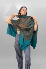 Ruana de lana merino tejida, teñida y estampada artesanalmente. Presenta un cuello plisado en fieltro de lana merino y un estampado con brillo en tonos dorados/bronce. El color principal es un degradado de gris oscuro a turquesa vibrante.