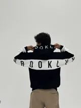 Canguro con capucha de corte boxy, color negro con una franja horizontal blanca que recorre la espalda y las mangas, presentando la palabra 'BROOKLYN' estampada en negro sobre la franja. La capucha también incluye detalles estampados.