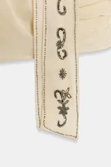 Camisa color crema o marfil, de tejido semitransparente, con escote pico cruzado y mangas largas con puños abotonados. Presenta un detalle de foulard incorporado en el frente, adornado con apliques de abalorios oscuros en forma de motivos florales y geométricos.
