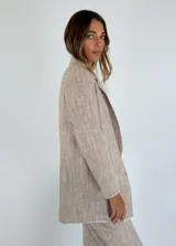 Blazer de lino y algodón en tono beige con textura a rayas verticales sutiles. Presenta solapas clásicas, un solo botón frontal marrón y un corte ligeramente holgado. Los bordes de las mangas y el ruedo tienen un acabado deshilachado.