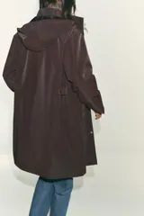 Chubasquero largo color marrón oscuro, con cuello subido y capucha ajustable con cordones. Tiene cierre frontal con cremallera oculta y botones a presión, y bolsillos delanteros con solapa y botón a presión oculto.