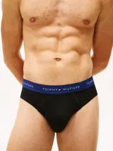 Pack de tres calzoncillos tipo slip de algodón elástico con cintura elástica personalizada con el logo y nombre de la marca Tommy Hilfiger. Cada unidad presenta una banda elástica de distinto color: verde oliva, marrón y azul marino, sobre una base de color negro.