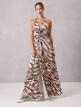Vestido largo de tejido satinado plisado con estampado animal print tipo cebra en tonos tierra y ocre. Presenta escote halter profundo en V con drapeado central y broche de pedrería, espalda descubierta y falda fluida con apertura frontal.