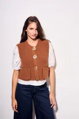 Chaleco tejido color beige con cuello redondo, sin mangas, con cierre frontal de cuatro botones dorados y dos bolsillos frontales.