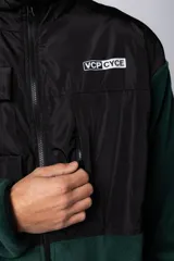 Campera de polar con recortes en tela técnica tipo sire en la parte superior frontal. Presenta cierre frontal completo, cuello alto y bolsillos funcionales en el pecho. Diseño bicolor en tonos verde oliva y negro con logo de la marca en el pecho.
