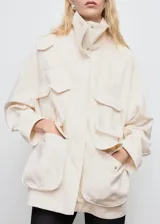 Anorak de estilo utilitario en color crudo, con cuello alto tipo embudo, cierre frontal con cremallera y botones a presión, y múltiples bolsillos de parche con solapa. Presenta un diseño oversize de manga larga.