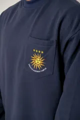 Buzo de cuello redondo color azul marino, con manga larga y corte holgado. Presenta un bolsillo frontal en el pecho con un bordado del sol de la bandera uruguaya, cuatro estrellas y la frase 'Heroicos sabremos cumplir'.