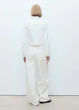 Chaqueta crop de denim blanco, confeccionada en algodón con un toque de elastano. Presenta cuello de solapa, cierre frontal con botones metálicos, bolsillos de parche con solapa en el pecho y manga larga con puños abotonados.