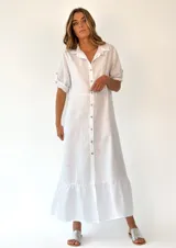 Vestido largo camisero de lino blanco, con cuello clásico, mangas cortas con presilla y botones, cierre frontal con botones y falda con volantes.