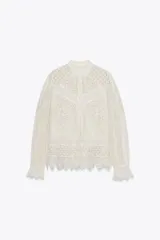 Blusa blanca de algodón con cuello redondo, manga larga con volante en el puño y detalle de bordados y aplicaciones de encaje en paneles geométricos. Cierre frontal con botones.