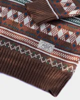 Buzo tejido de punto con diseño jacquard de motivos geométricos y nórdicos en tonos marrón, verde, blanco y azul. Presenta cuello redondo, puños y ruedo acanalados, y etiqueta de la marca Denali en el frente.