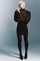 Vestido corto de color marrón con diseño combinado. Presenta un cuerpo superior fluido con cuello subido, lazada frontal ajustable y mangas largas abullonadas con hombreras y puños ajustados. La parte inferior es entallada con tejido elástico a tono, espalda con escote fluido y cierre lateral con cremallera oculta.