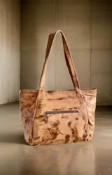 Cartera tipo shopper de cuero suave color marrón terra, con diseño pintado a mano que incluye salpicaduras y la inscripción de la marca. Cuenta con dos asas largas para llevar al hombro, cierre principal, forro interior con bolsillo y un dije metálico colgante.