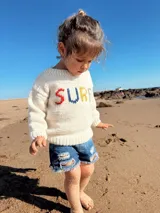 Sweater tejido a mano en lana merino color crudo con cuello redondo y mangas largas. Presenta la palabra "SURF" bordada en el frente con letras de colores.