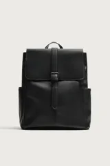Mochila de diseño minimalista en color negro, confeccionada en material sintético con acabado liso. Presenta una solapa frontal con cierre de hebilla decorativa y sistema de imán, compartimento principal con cremallera, bolsillos laterales externos, asa superior de mano y correas de hombro ajustables y acolchadas.