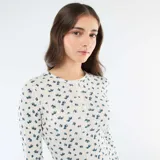 Camiseta de manga larga confeccionada en tejido de punto calado, con estampado de pequeñas flores azules sobre fondo blanco. Presenta un diseño ajustado al cuerpo, cuello redondo y una pequeña abertura frontal con botones.