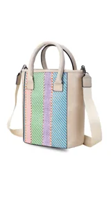 Matera de diseño vertical con paneles laterales y asas en material símil cuero color beige. El panel frontal presenta un tejido artesanal con rayas diagonales multicolores en tonos verde, lila, rosa y azul. Incluye correa de hombro ajustable en color crema.