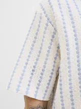 Camisa de manga corta estilo resort, confeccionada en tejido jersey de punto con diseño de rayas verticales en tonos crudo y azul. Presenta cuello camisero y cierre frontal con botones.