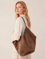 Cartera tipo hobo de gran capacidad confeccionada en símil gamuza color marrón. Presenta un diseño relajado y flexible con base reforzada y ribetes de cuerina a juego. Cuenta con una sola asa de hombro integrada a la estructura y un compartimento interior independiente.