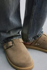 Zueco tipo mule de hombre, color beige topo, fabricado en piel con acabado serraje. Presenta una tira con hebilla metálica cuadrada en el empeine y suela gruesa de goma en color natural contrastante.