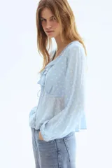 Blusa celeste con estampado de lunares blancos, escote en V pronunciado con volados, mangas largas abullonadas con puños elásticos y lazo ajustable en el frente.