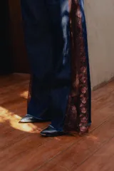 Pantalón de jean estilo wide leg, confeccionado en denim rígido de color azul oscuro. Presenta paneles laterales de encaje con diseño floral en tono rojizo y terminaciones deshilachadas en el ruedo.