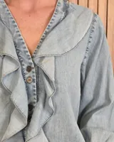 Camisa de denim de algodón ligero, con cuello en V adornado con grandes volados frontales y cierre de botones. Presenta mangas largas con puños elásticos fruncidos.