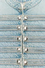 Chaleco de denim color celeste con cuello subido, sin mangas, con detalle de aplicaciones horizontales y botones metálicos en el frente. Cuenta con cierre frontal con ganchos metálicos ocultos.