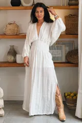 Vestido largo blanco de estilo bohemio, con escote en V profundo, mangas largas abullonadas y cintura fruncida elastizada. Presenta un diseño fluido con sutiles rayas verticales tejidas en la tela y una abertura lateral en la falda.