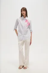 Camisa de algodón con rayas verticales azules y blancas, corte ancho y manga larga con lazos ajustables en los puños. Presenta un bordado de dos palmeras en color rosa brillante en el área del pecho.