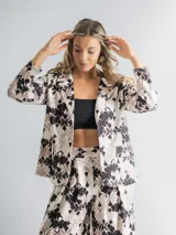 Conjunto de dos piezas compuesto por un blazer de corte recto con solapas y un pantalón de tiro alto y pierna ancha. Ambas prendas están confeccionadas en tela con estampado floral en tonos neutros. El blazer incluye un aplique floral desmontable.