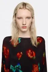 Vestido midi de corte ajustado con falda superpuesta de tul, escote barco y mangas largas tipo murciélago. Presenta un diseño estampado con motivos florales en tonos rojos y verdes sobre fondo negro, con detalles fruncidos en la cintura.