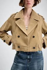 Trench corto cruzado de piel de ante color tostado, con cuello de solapa, bolsillos delanteros de vivo y manga larga con trabillas en los puños.