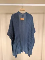 Ruana tejida de algodón con hilo lurex en punto jersey, color azul, con diseño amplio y detalle de etiqueta de cuerina en la parte posterior.