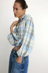 Camisa de cuello solapa y escote en pico, con diseño de cuadros en tonos azules, blancos y verdes. Presenta cierre frontal cruzado con lazada lateral y mangas largas con puños elásticos.
