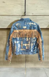 Campera de jean azul intervenida con detalles en tono dorado pálido, con flecos de cuero beige en la espalda y las mangas, decorados con tachas metálicas.