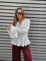 Blusa blanca de textura arrugada, con escote en V y lazos para anudar. Presenta mangas largas con puños acampanados y un diseño con volado en el ruedo.