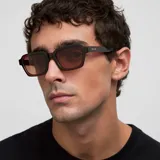 Lentes de sol unisex modelo Adisa, con montura gruesa de forma cuadrada y patillas robustas. El color de la montura es marrón oscuro (maroon) y los lentes son de color marrón.