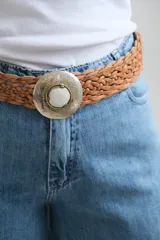 Cinto trenzado de cuero color camel con hebilla metálica circular de estilo artesanal, decorada con un detalle central en piedra clara.