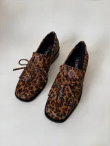 Mocasín de cuero marrón con textura granulada, diseño de horma cuadrada, detalle de flecos y lazo decorativo en el empeine, montado sobre una suela y taco bajo de color negro.