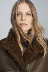 Chaqueta tipo bomber de cuello alto y manga larga, confeccionada en material con efecto cuero envejecido y forro interior de pelo sintético. Presenta cierre frontal con botones, bolsillos delanteros y bajo ajustable con cordones.