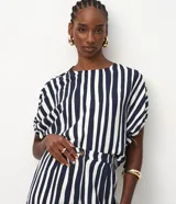 Blusa de viscosa con estampado de rayas verticales blancas y azul marino. Tiene cuello redondo, mangas cortas con detalle de lazo ajustable en los laterales y cierre de botón en la espalda.